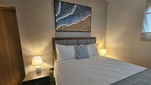Un letto rifatto con lenzuola bianche e una coperta grigia, affiancato da due comodini con lampade, in una stanza con un grande quadro astratto sopra il letto.