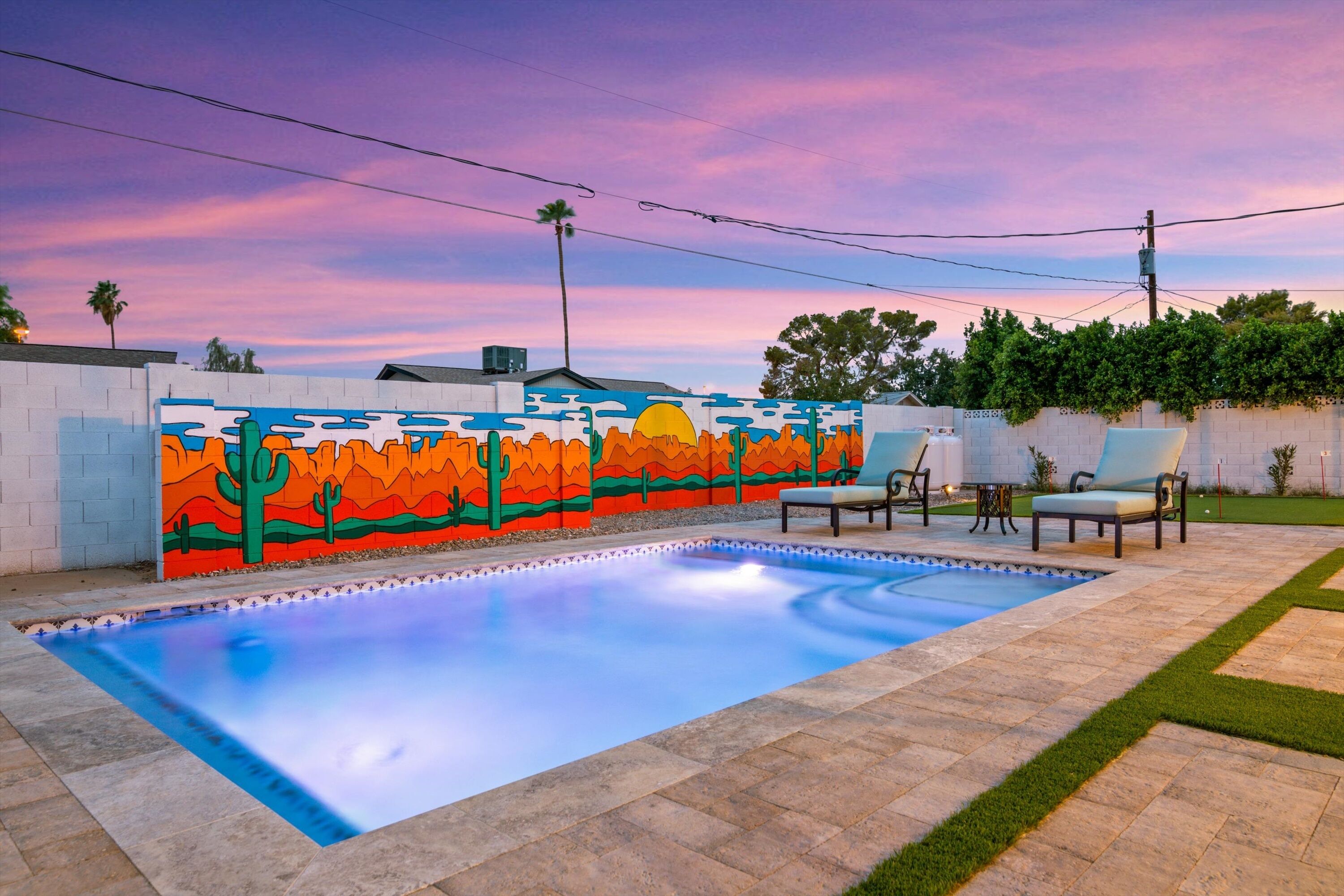 A rectangular swimming pool with a colorful mural on the adjacent wall, surrounded by a paved patio with chairs and a small lawn area.