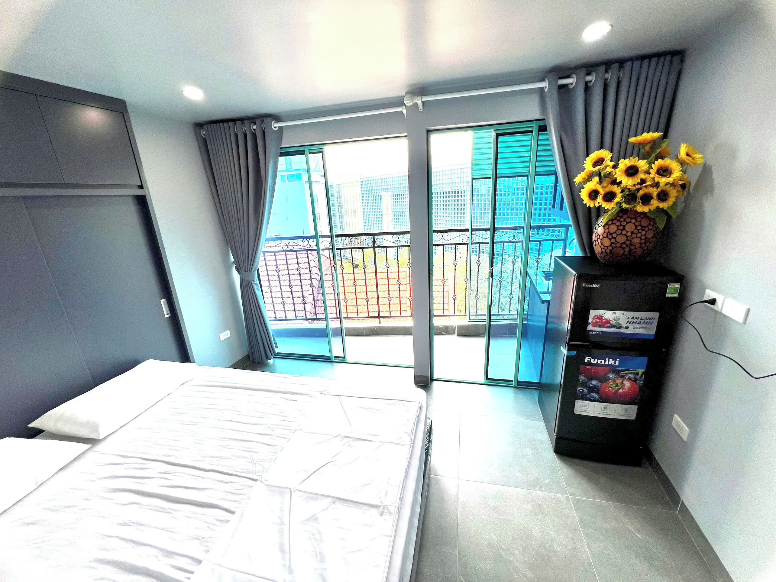 A hotel room with a bed, a minibar, a view of the outside, and a vase with sunflowers.