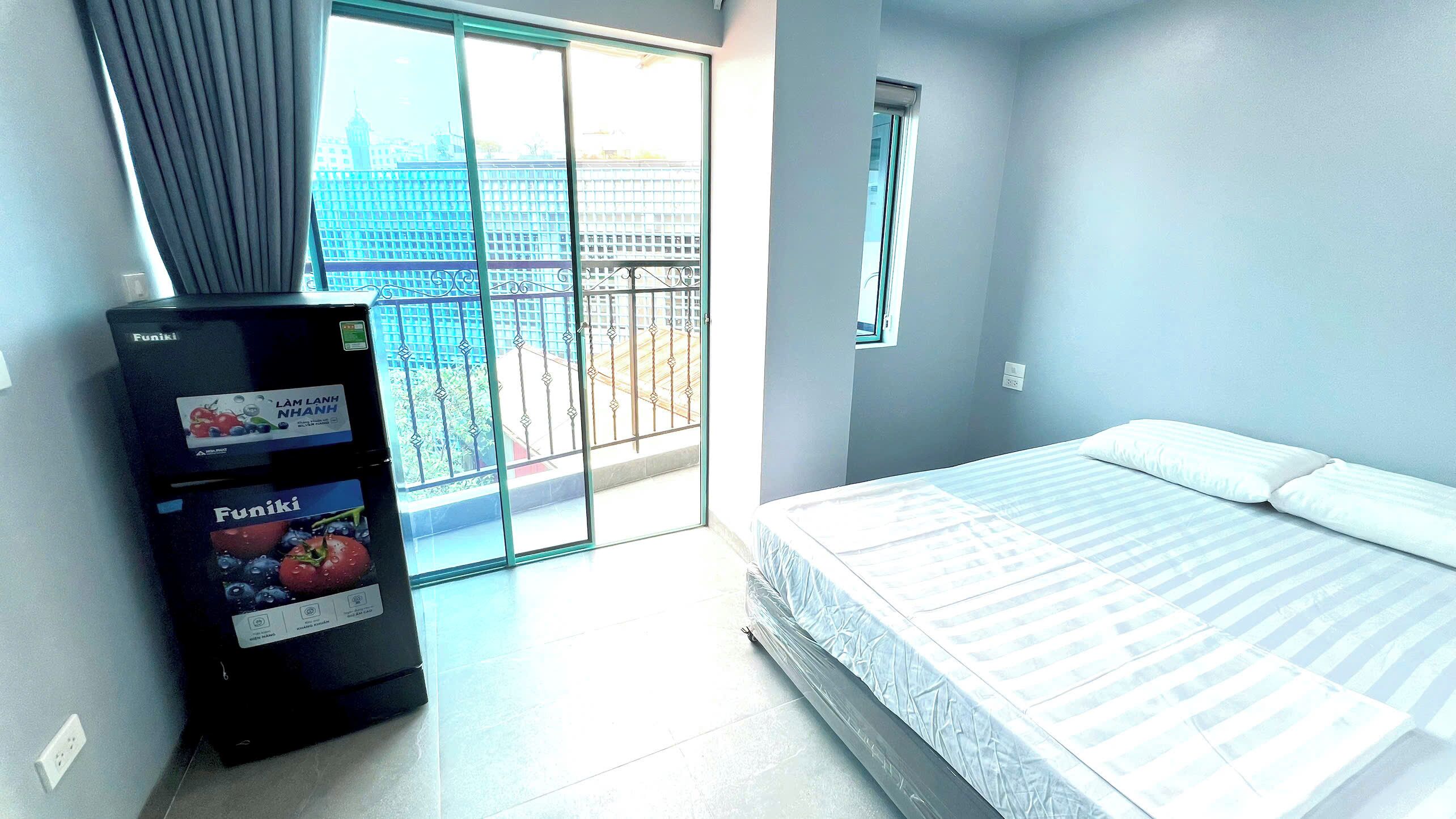 A hotel room with a bed, a vending machine, a window with a view of a cityscape, and a balcony with a railing.