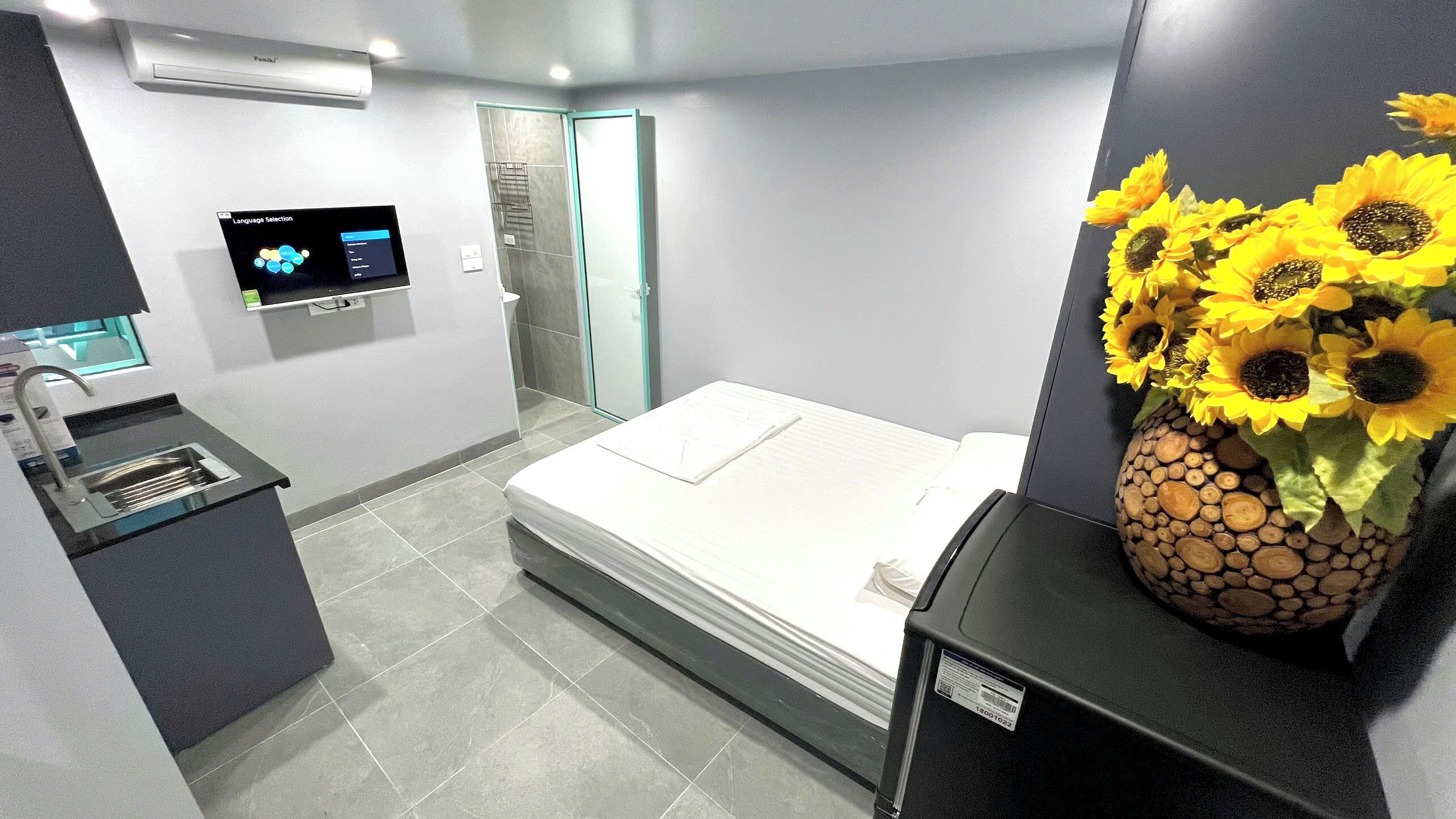 A modern hotel room with a bed, a small sink, a shower, and a vase of sunflowers.