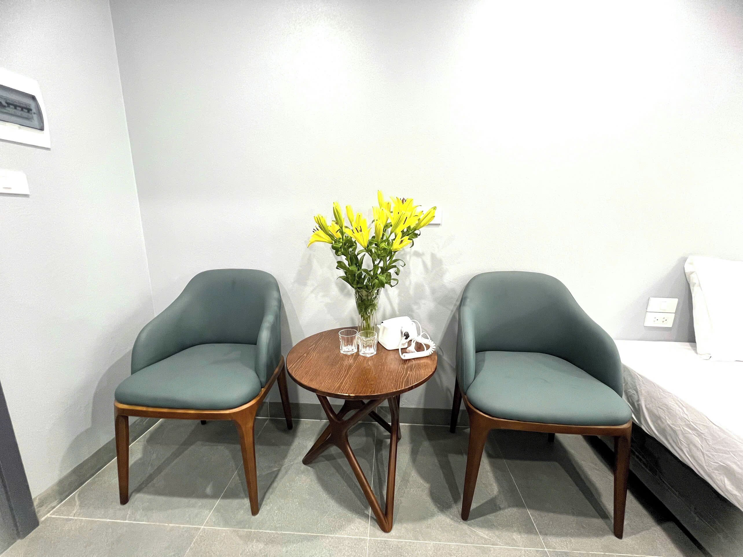 Una habitación con dos sillas verdes, una pequeña mesa de madera con un jarrón de flores amarillas, un teléfono blanco y una cama con ropa de cama blanca.