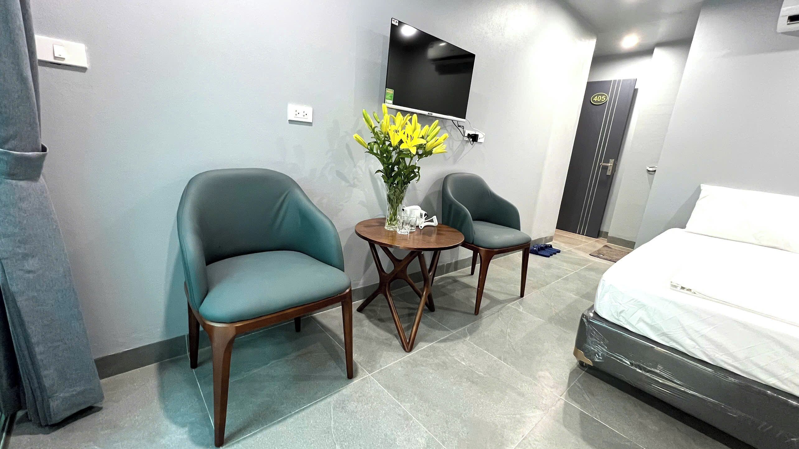 A hotel room with two chairs, a small table with a vase of flowers, a bed, a TV mounted on the wall, and a door with a number sign.