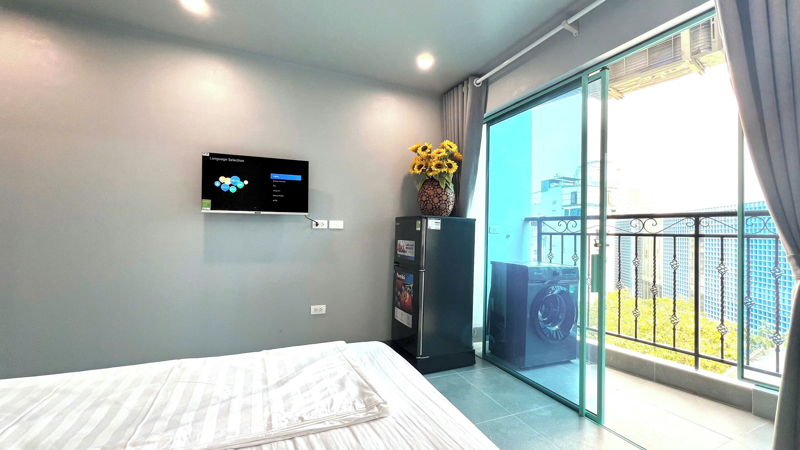 A modern bedroom with a bed, a mounted TV, a refrigerator, and a balcony with a view of buildings.