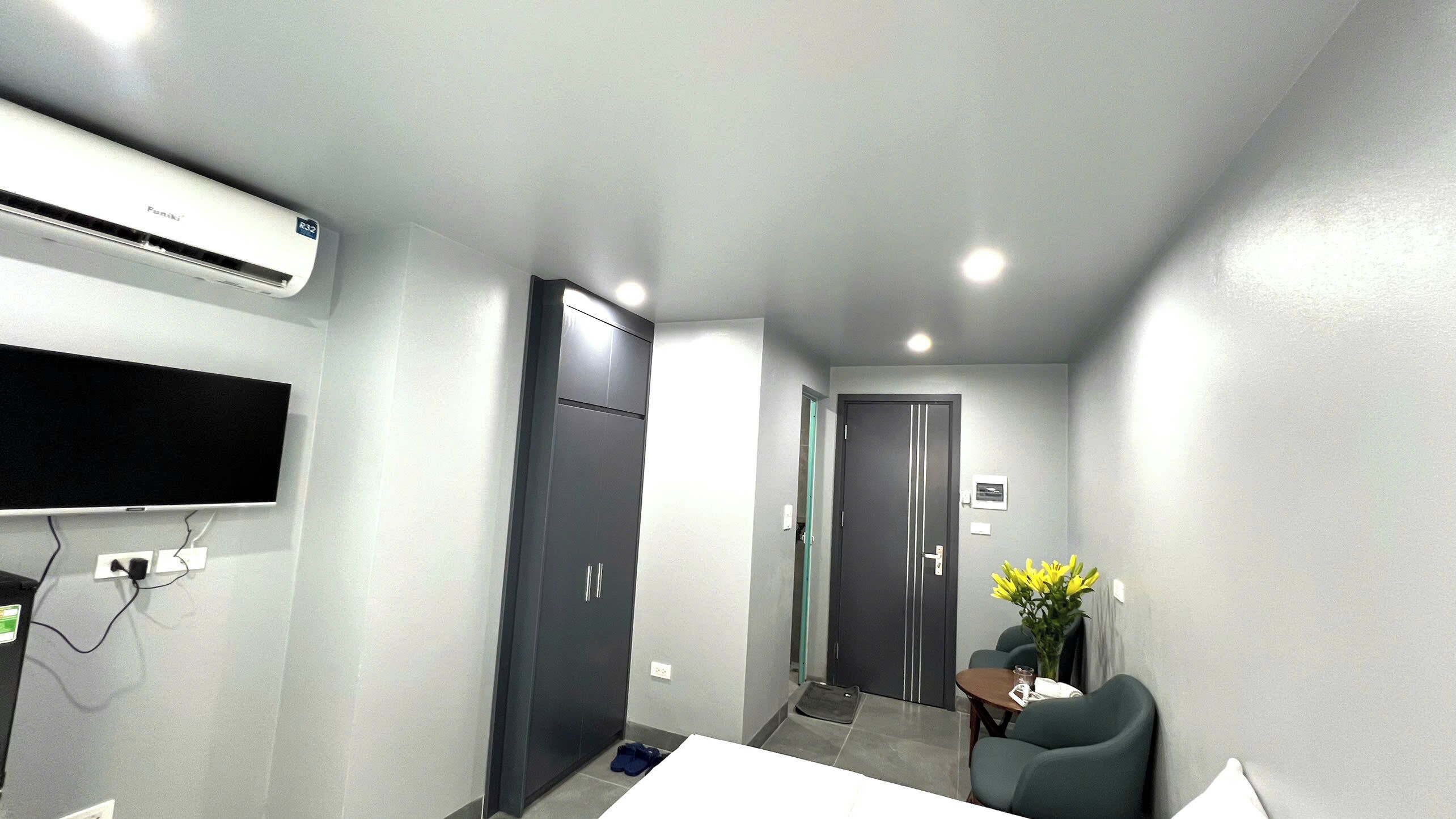 A hotel room with a flat-screen TV mounted on the wall, a desk with a chair, a small table with a vase of flowers, and a door leading to another room.