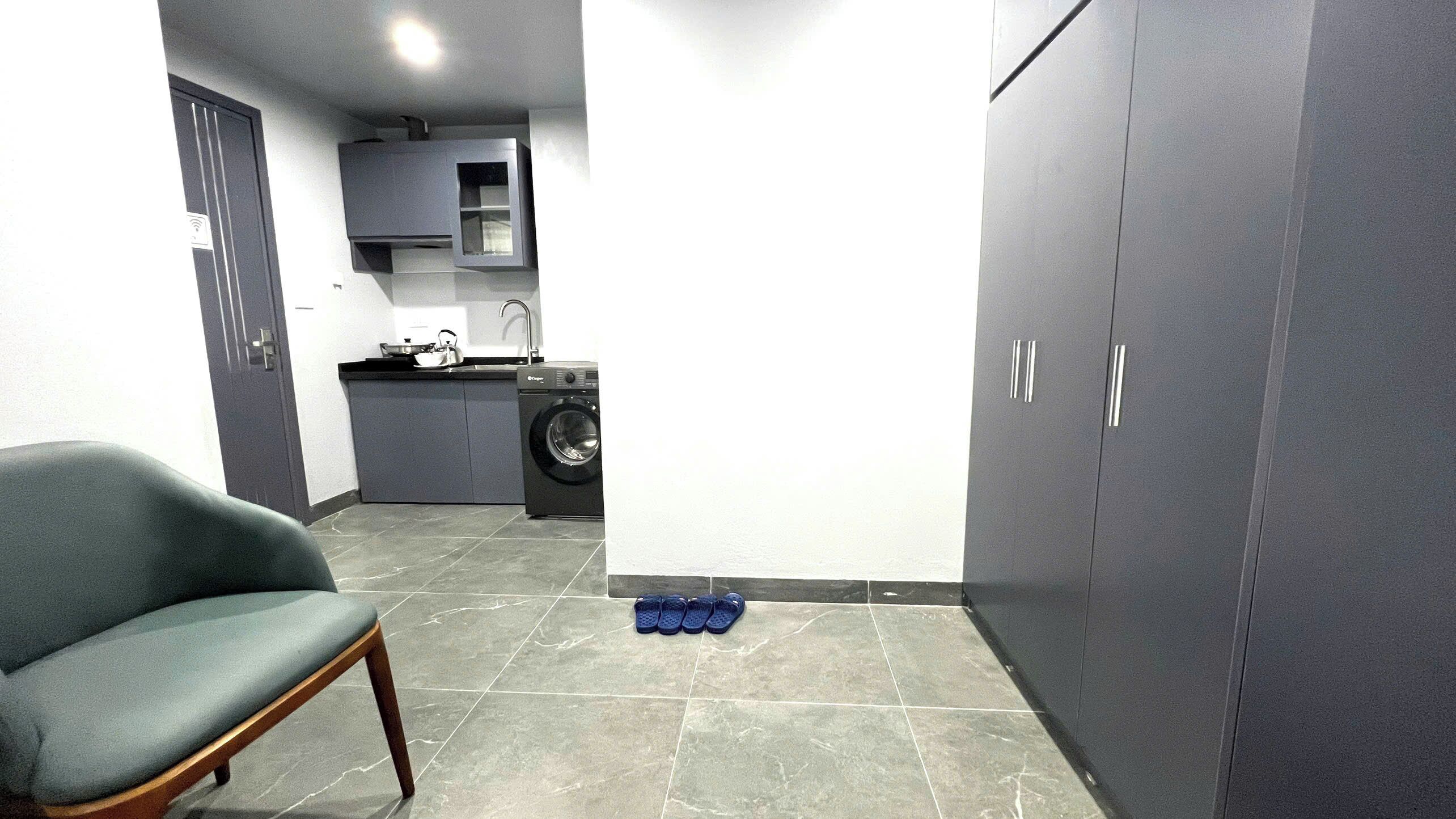 Ett kompakt bostadsutrymme med en grå soffa, en tvättmaskin och ett litet köksutrymme.