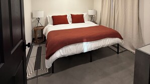 Ein ordentlich eingerichtetes Schlafzimmer mit einem Bett, zwei Nachttischlampen, einem Nachttisch, einem gemusterten Teppich und einer dunkelholzigen Tür.