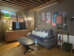 A living room with a sofa, a coffee table, a TV, and artwork on the walls. - Cozy apartment in a historic medieval town. (Chalon-sur-Saône)