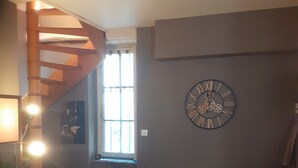 A room with a wooden staircase, a large wall clock, a small table with a chair, and a window with curtains.