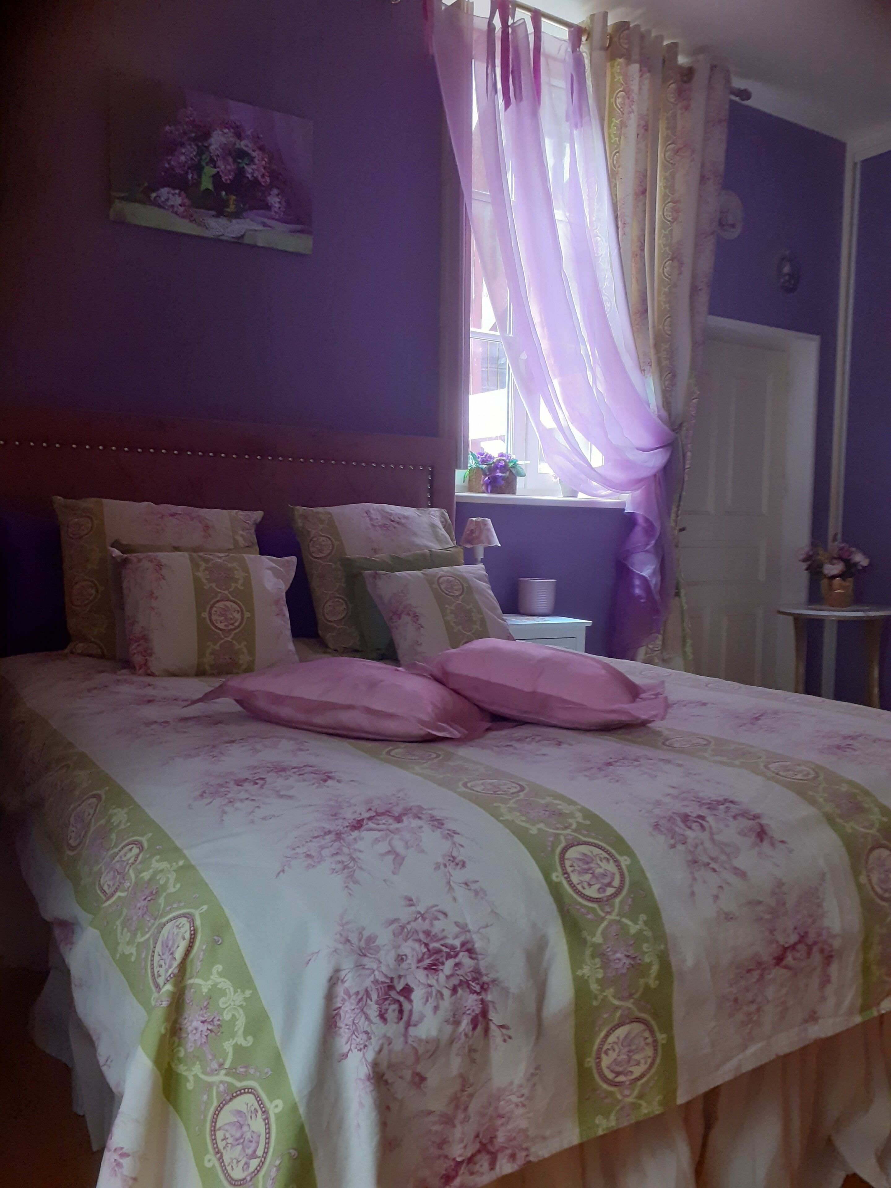Une chambre à coucher avec un lit, des oreillers, une table de chevet, une lampe et une fenêtre avec des rideaux.