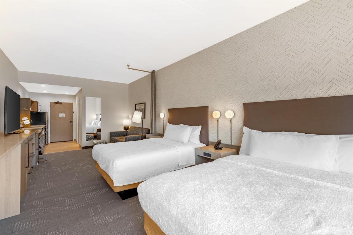 A hotel room with two beds, a desk with a flat-screen TV, a desk lamp, and a wall-mounted light fixture.