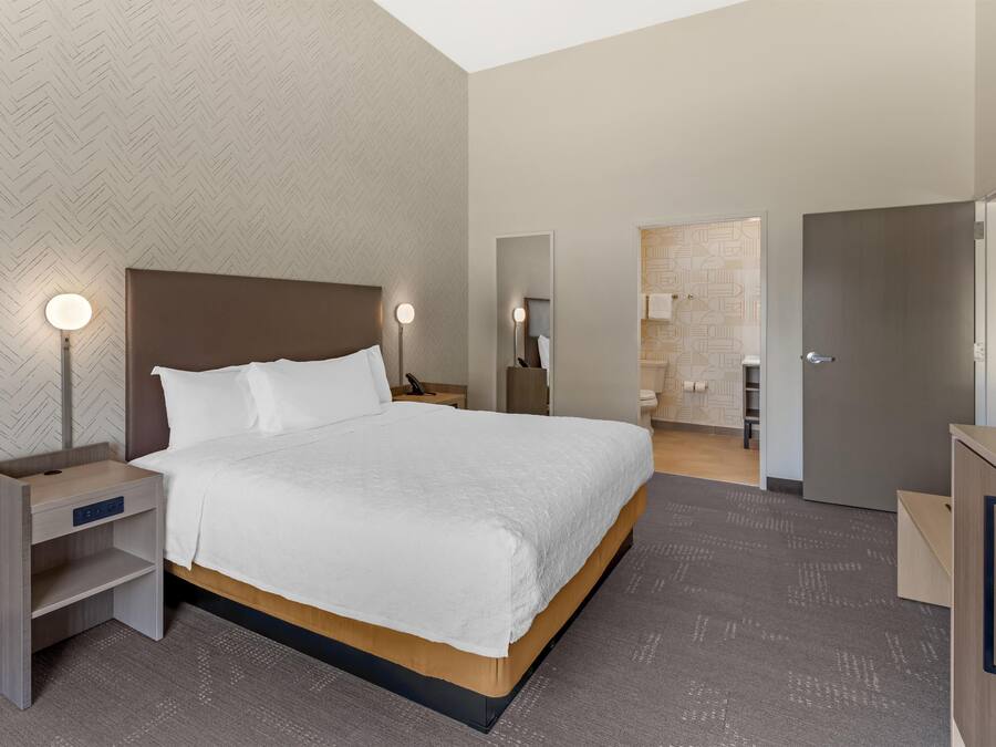 A modern hotel room with a large bed, bedside tables, a nightstand, a wardrobe, and a bathroom visible through an open door.