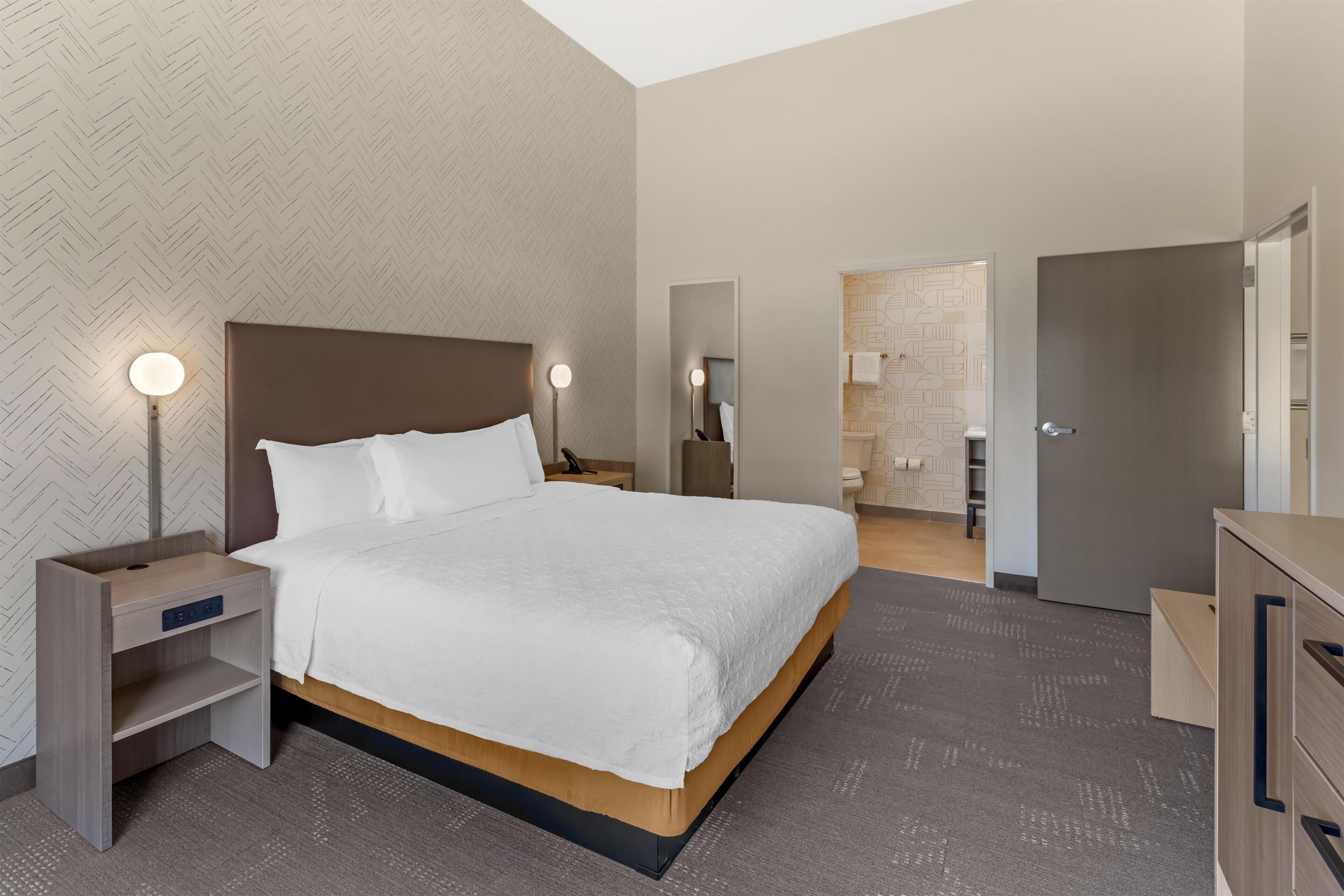 A modern hotel room with a large bed, bedside tables with lamps, a wardrobe, and a bathroom visible through an open door.