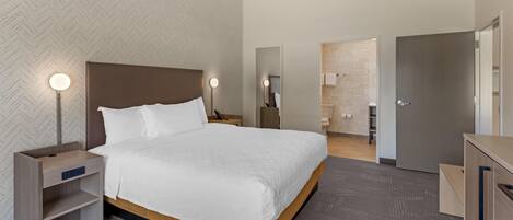 A modern hotel room with a large bed, bedside tables with lamps, a wardrobe, and a bathroom visible through an open door.