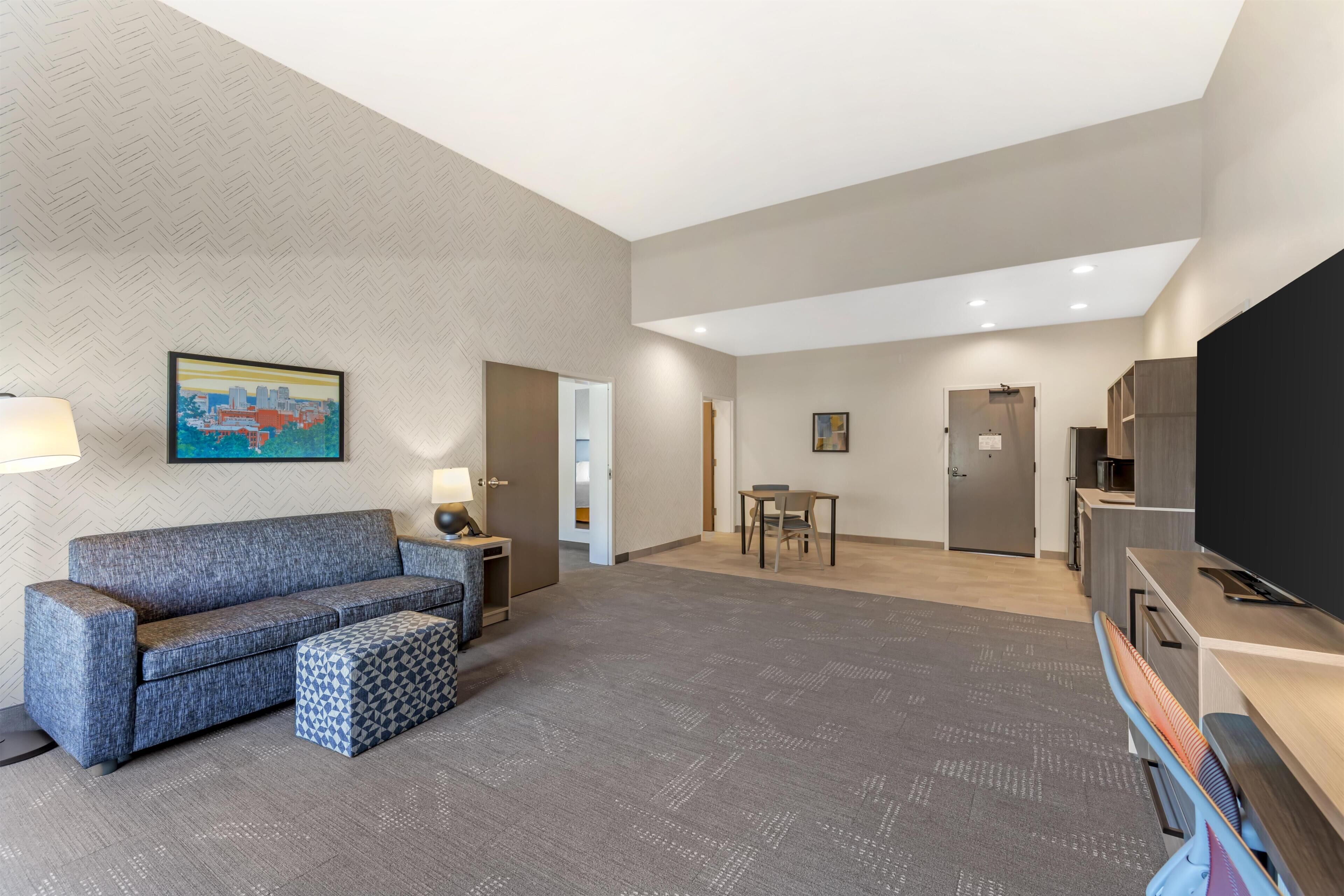 A modern hotel room with a sofa, ottoman, flat-screen TV, kitchenette, and a cityscape painting on the wall.