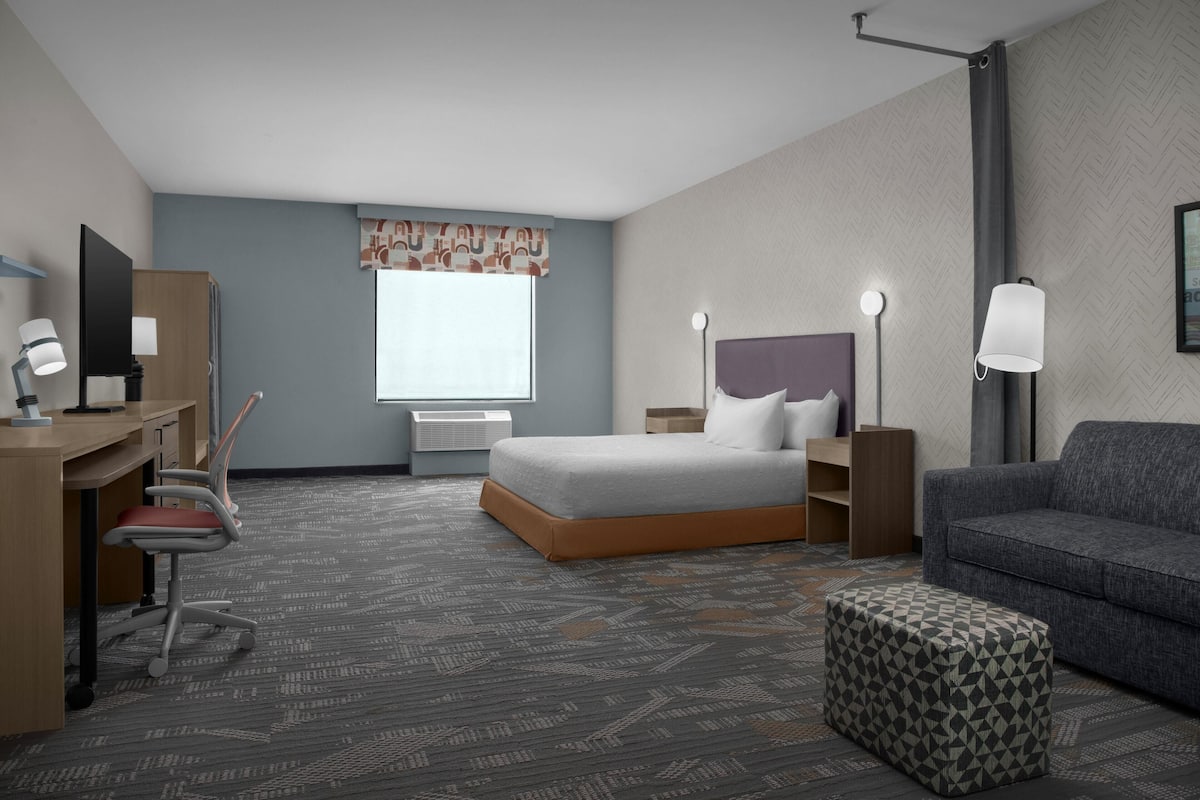 Ein Hotelzimmer mit Bett, Schreibtisch, Stuhl, Sofa, Hocker, Nachttischlampen und einem fenster mit Vorhängen.