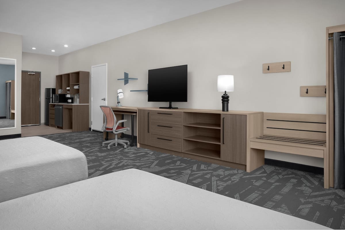 Ein Hotelzimmer mit zwei Betten, einem Schreibtisch, einem Fernseher, einem Sessel, einer Lampe und einer Küchenzeile.