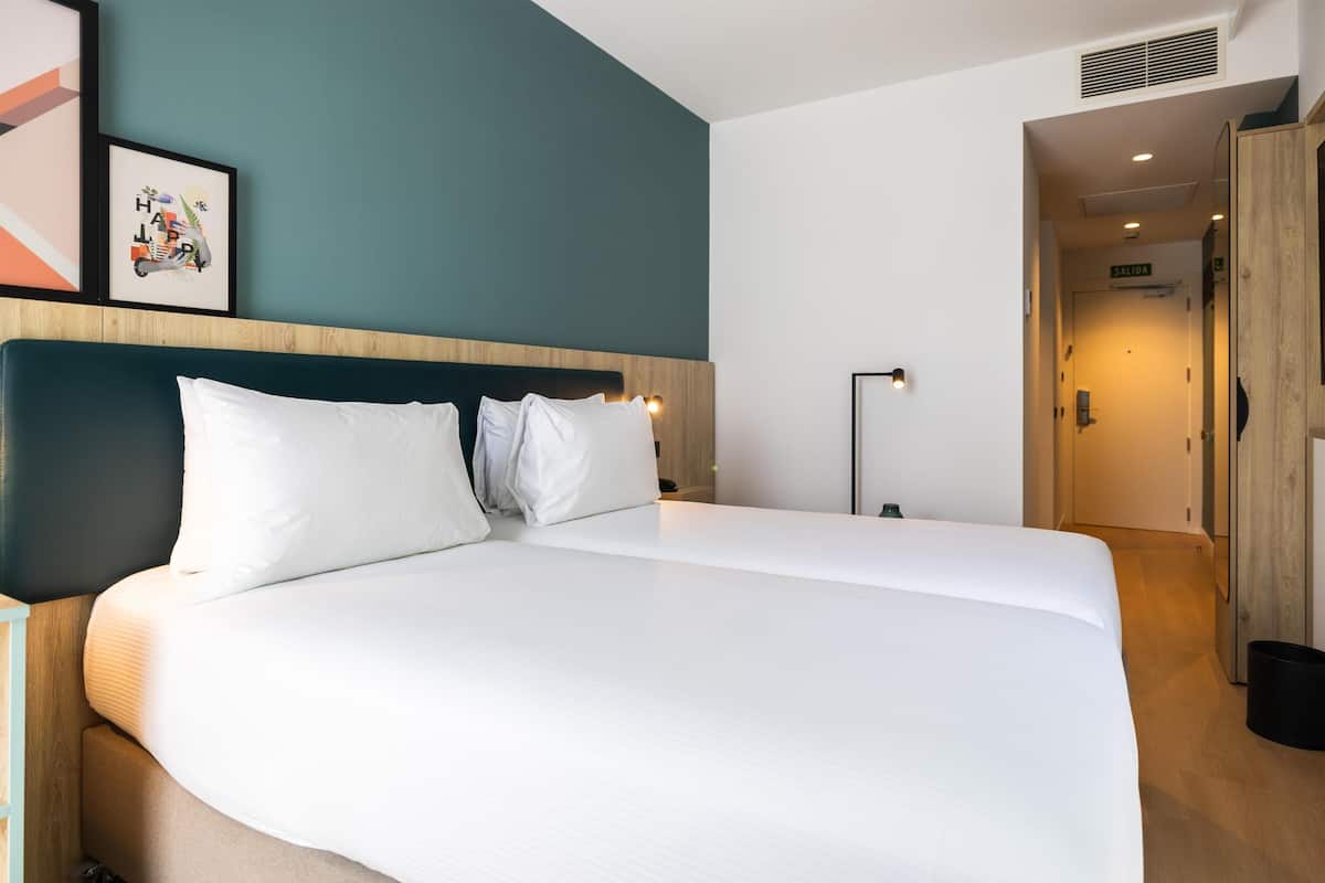 Una cama bien hecha en una habitación de hotel con una pared de acento color turquesa, un cuadro enmarcado y un pasillo que conduce a la salida.