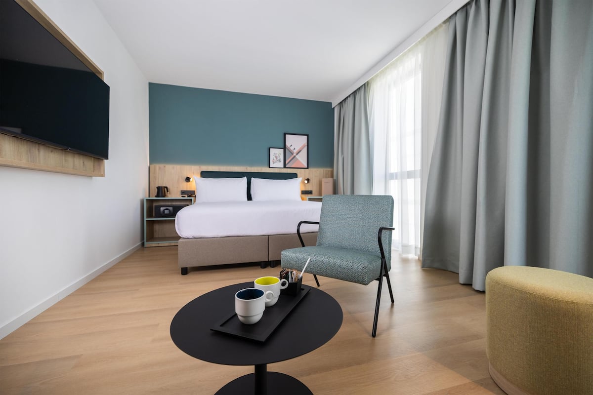Habitación de hotel con cama, una silla, una mesa con tazas, televisión y ventana con cortinas.