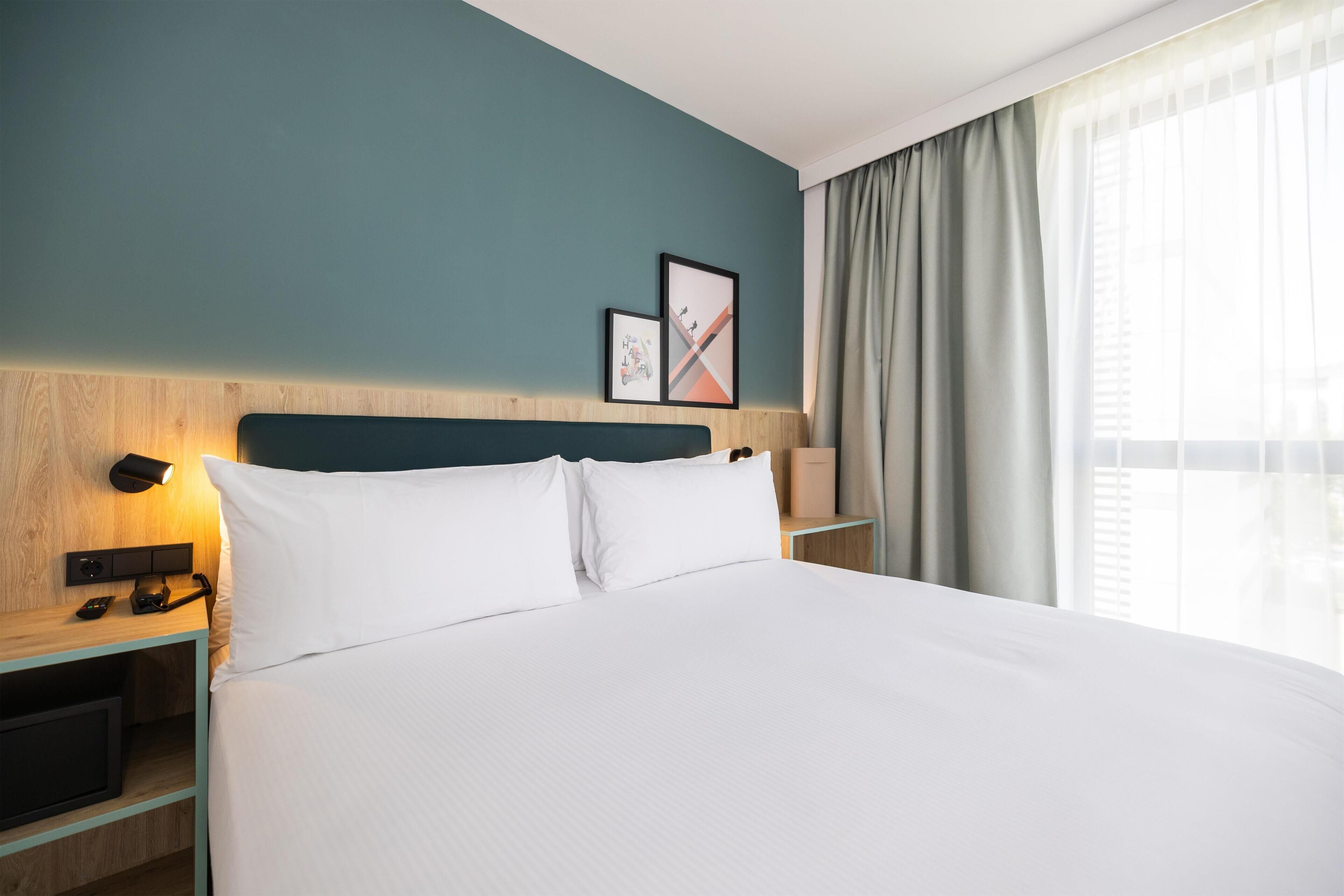 Ein Hotelzimmer mit einem großen Bett, einem hölzernen Kopfteil, einem Nachttisch mit Lampe und einem Fenster mit Vorhängen.