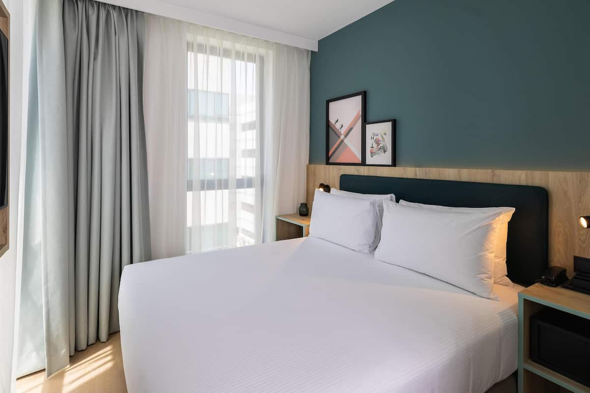 Habitación de hotel con una cama grande, cabecera de madera, mesita de noche y ventana con cortinas de gasa.