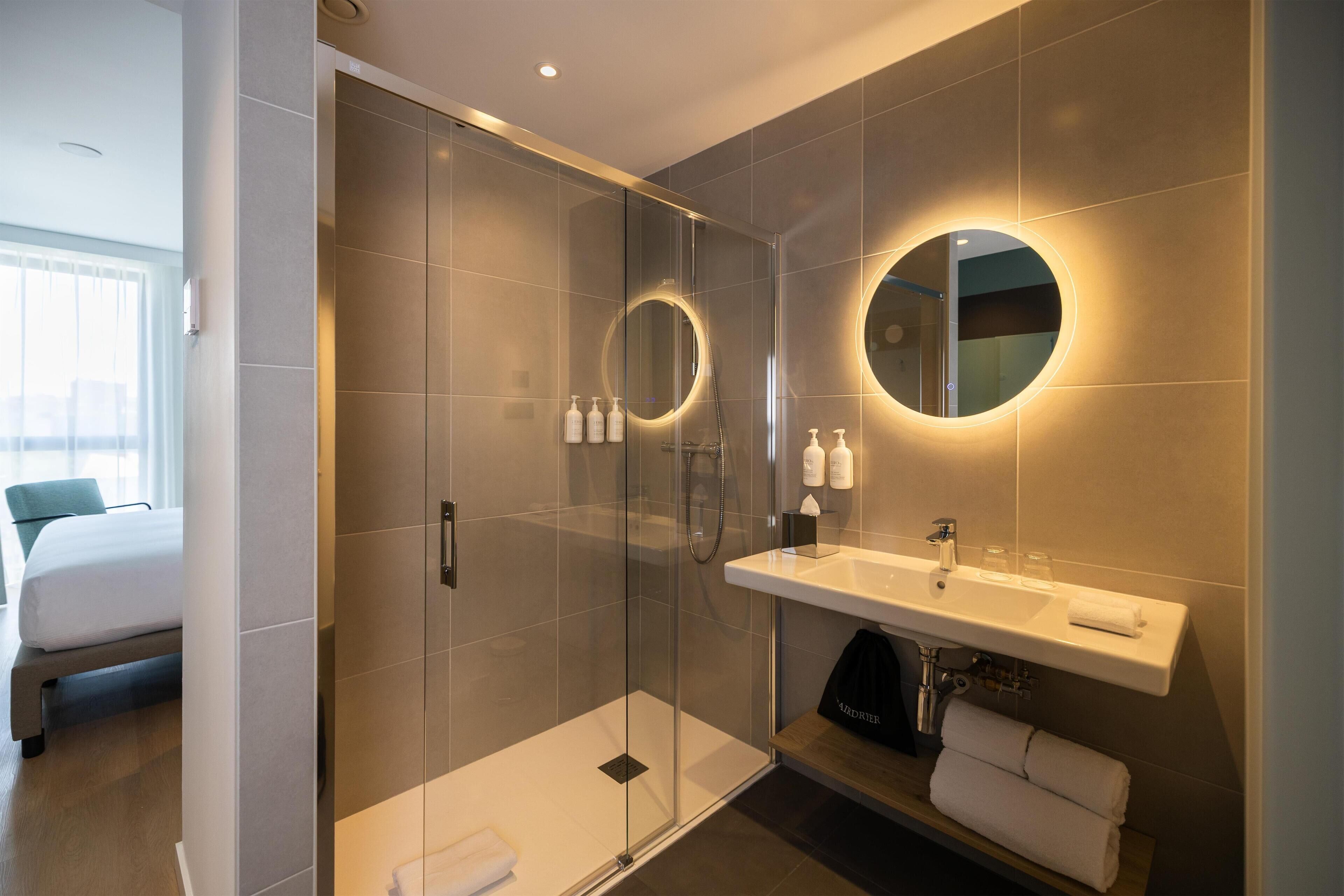 A modern bathroom with a glass-enclosed shower, a round mirror with a light, a white sink, and a bed in the adjacent room.