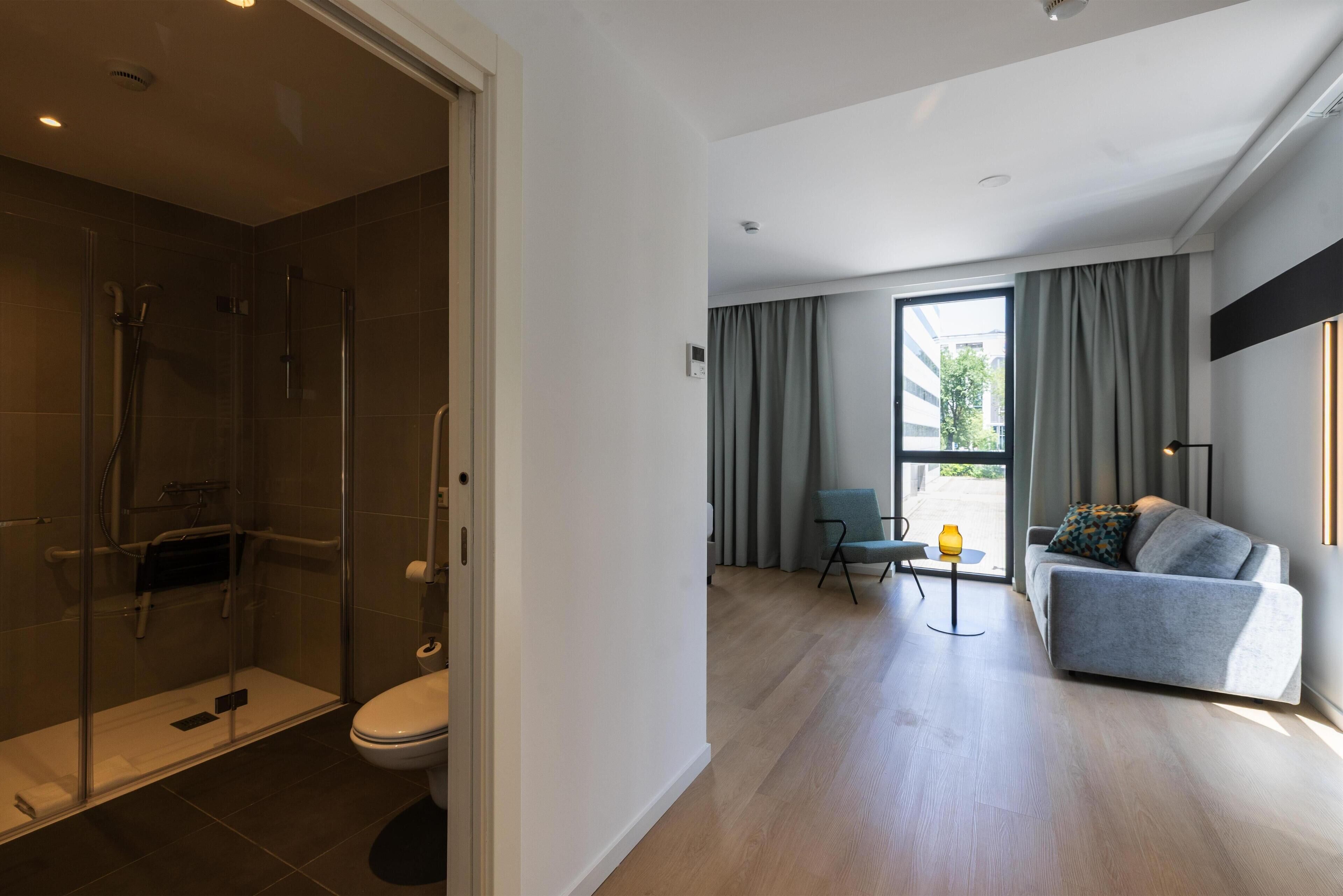 A modern bathroom with a glass shower enclosure, a toilet, and a small table with a lamp.