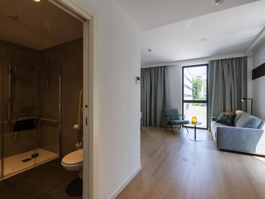 A modern bathroom with a glass shower enclosure, a toilet, and a small table with a lamp.