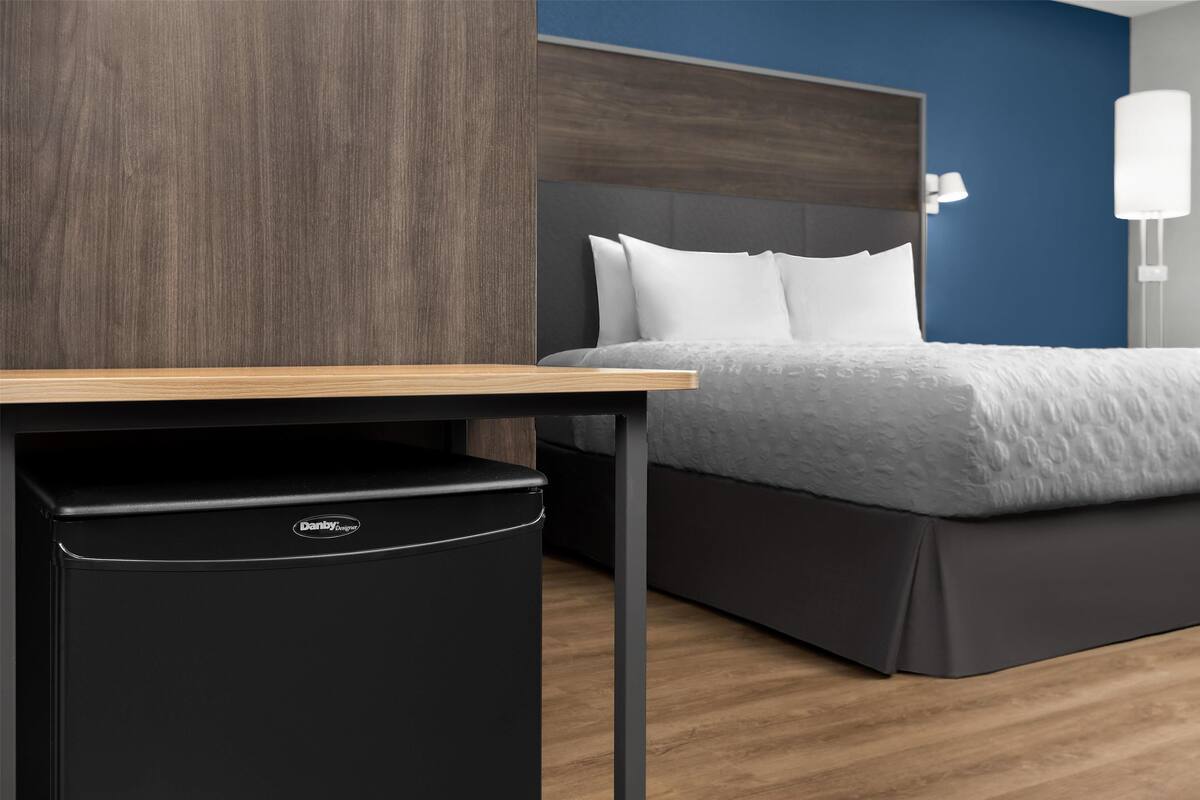 Habitación de hotel con un escritorio de madera, un cesto de basura negro, una cama con ropa blanca y una pared de acento azul.