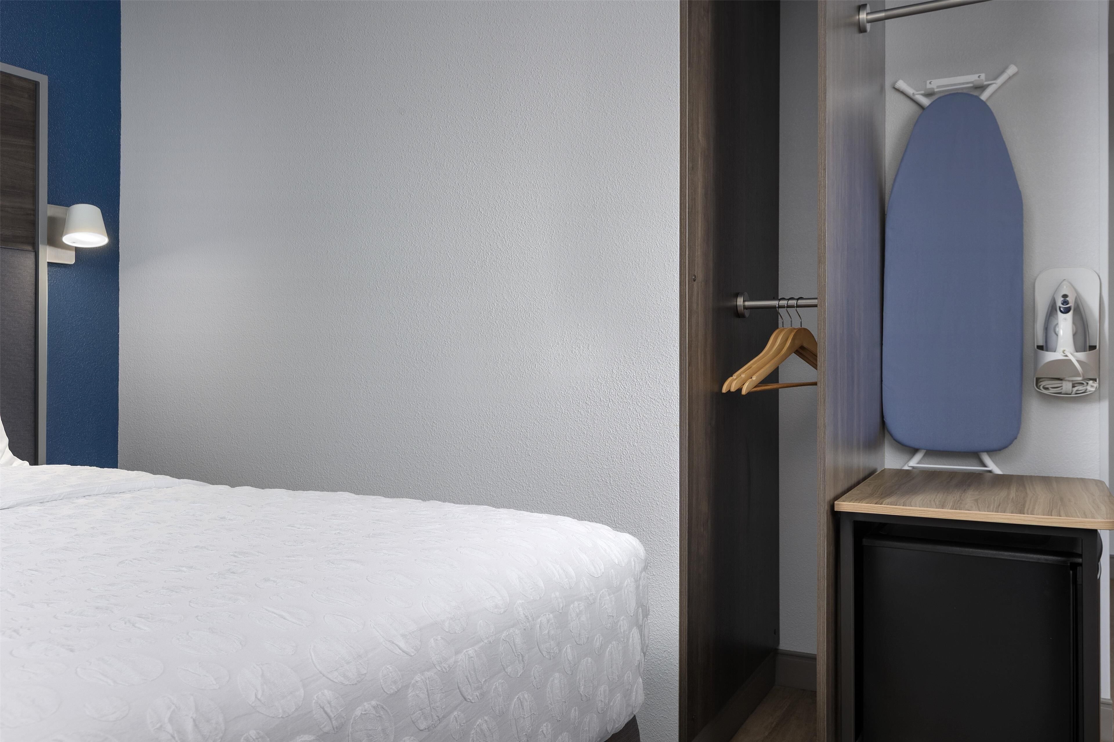 Een hotelkamer met een opgemaakt bed, een klein houten nachtkastje, een wandplank met een strijkplank en een handdoekrek.