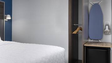 Een hotelkamer met een opgemaakt bed, een klein houten nachtkastje, een wandplank met een strijkplank en een handdoekrek.