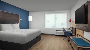 A modern hotel room with a large bed, a desk, a chair, a TV, and a window with blinds.