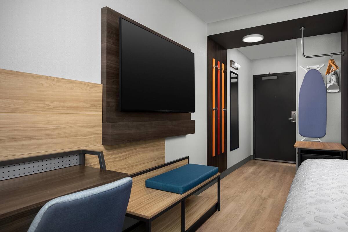 Una habitación de hotel moderna con un escritorio de madera, un banco acolchado azul, un televisor de pantalla plana, un armario y una cama con edredón estampado.