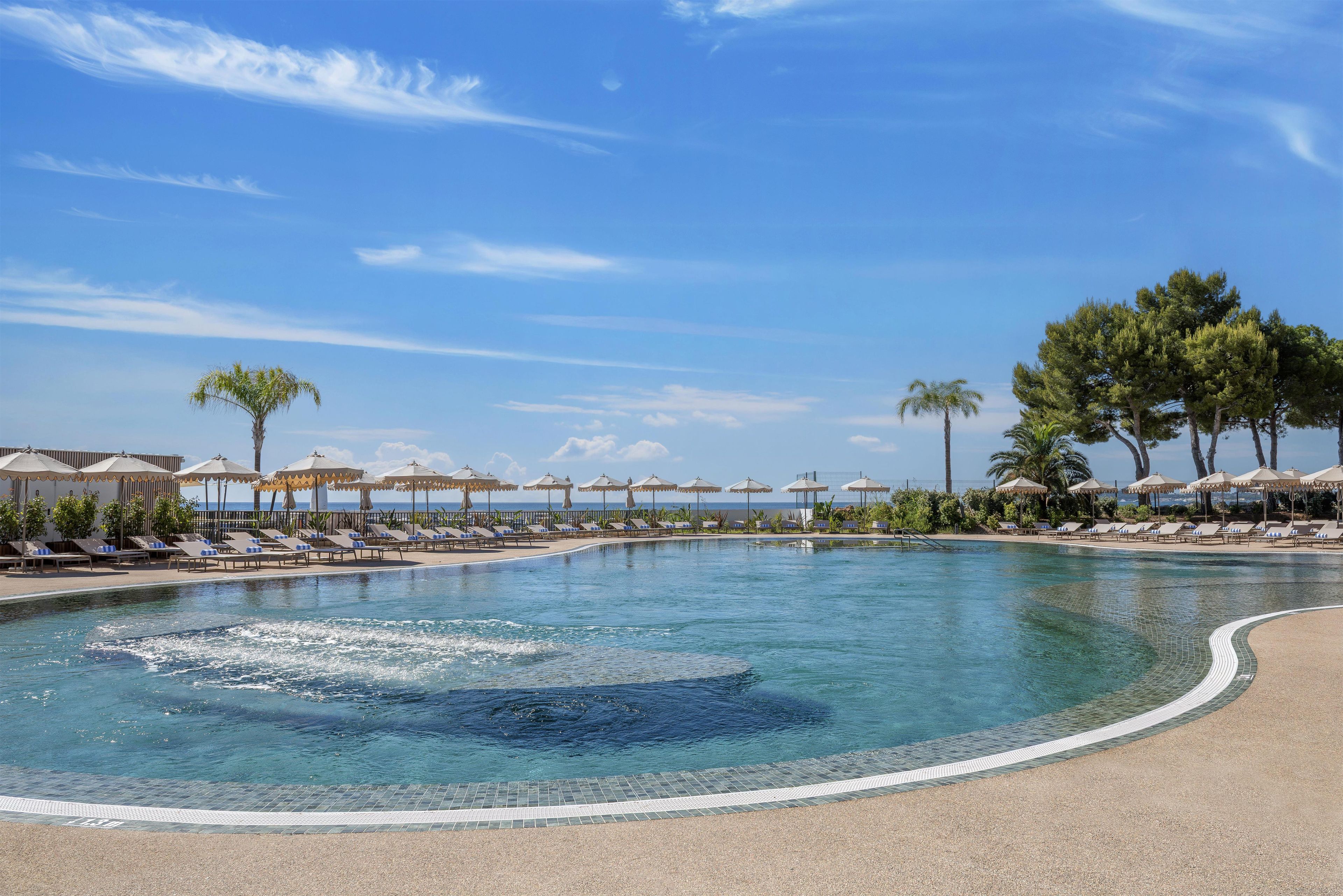 A pool with lounge chairs and umbrellas, surrounded by trees and a clear sky.