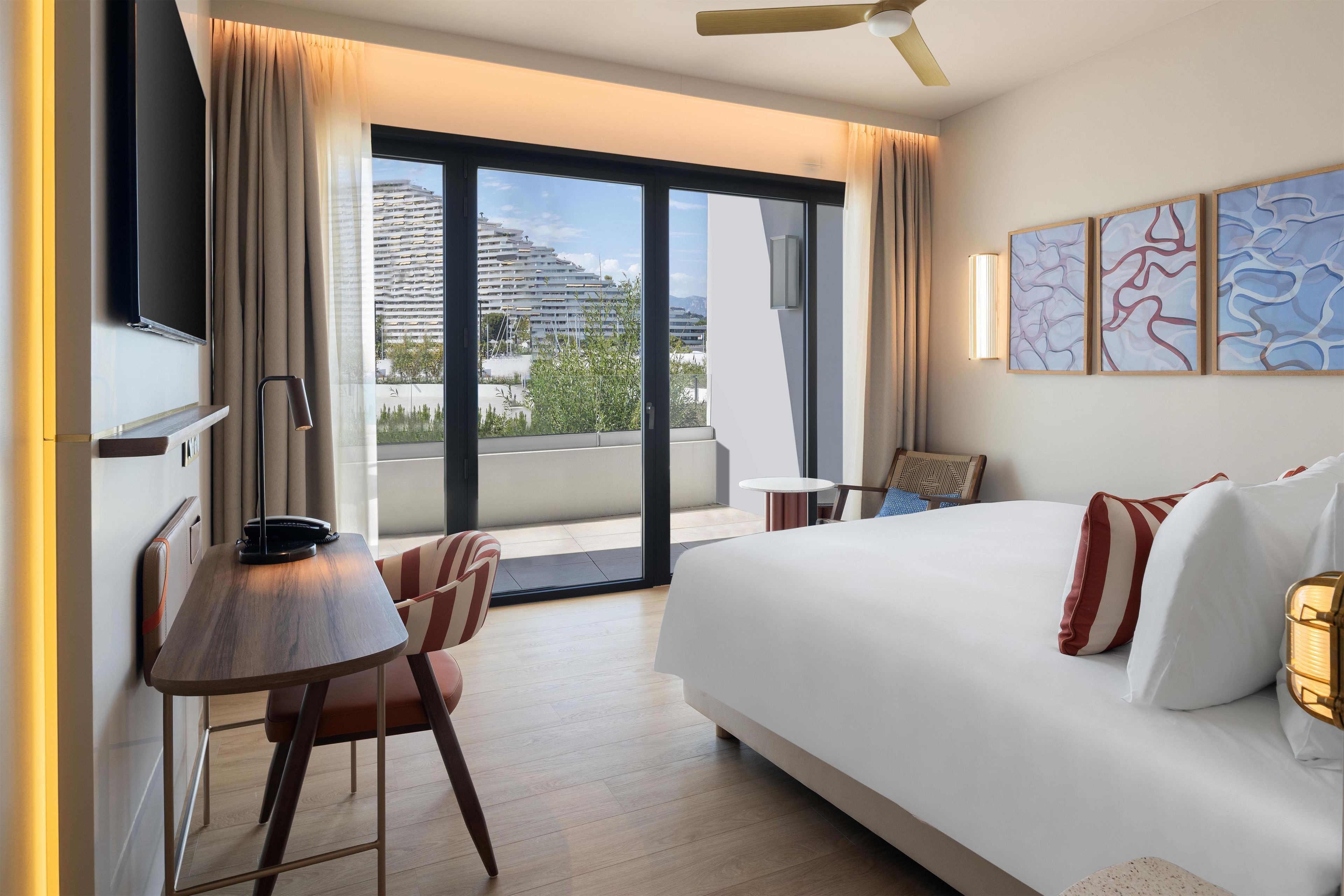 A modern hotel room with a large bed, a desk, a chair, a ceiling fan, and a view of a building through the window.