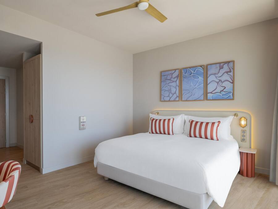 Quarto de hotel moderno com uma cama grande, um ventilador de teto, uma mesa de cabeceira com abajur e arte abstrata na parede.