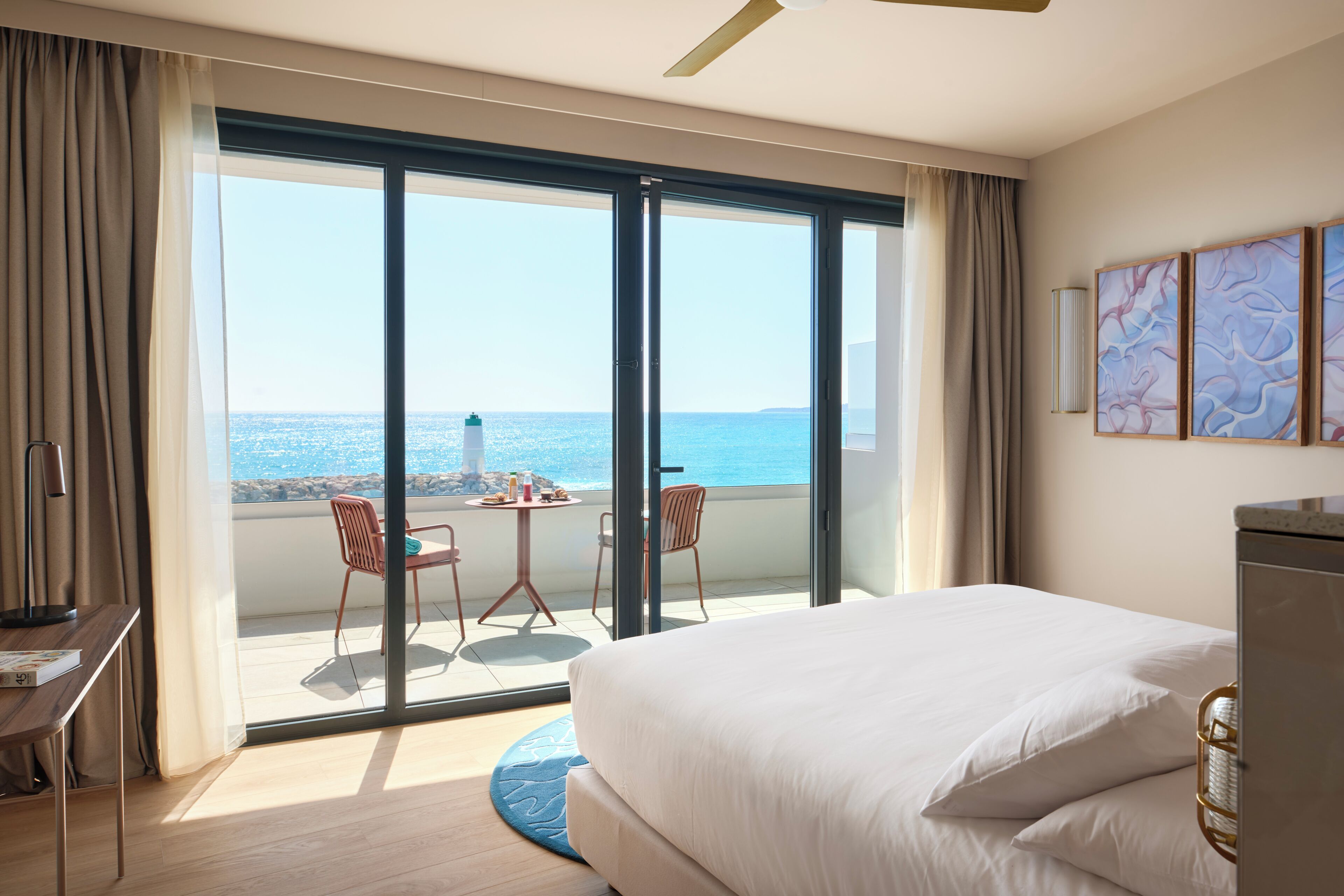 A hotel room with a large bed, a desk, a chair, a small table, and a view of the sea through a sliding glass door.