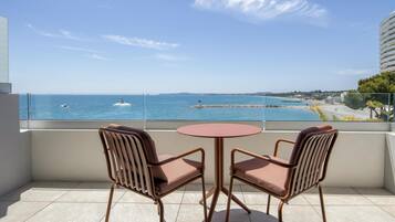 A balcony with two chairs and a table, overlooking a calm sea with a pier and distant shoreline.