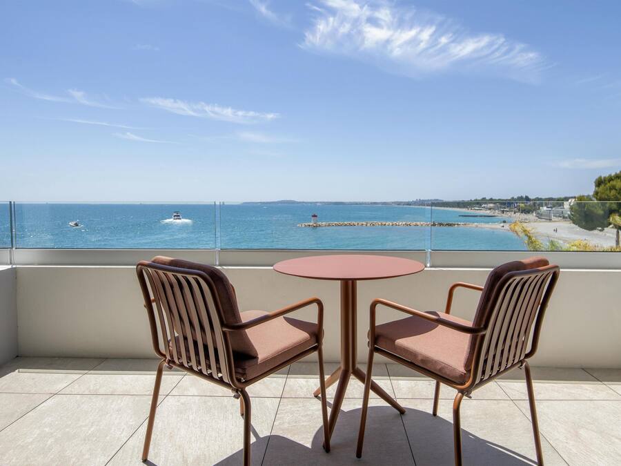 A balcony with two chairs and a table, overlooking a calm sea with a pier and distant shoreline.