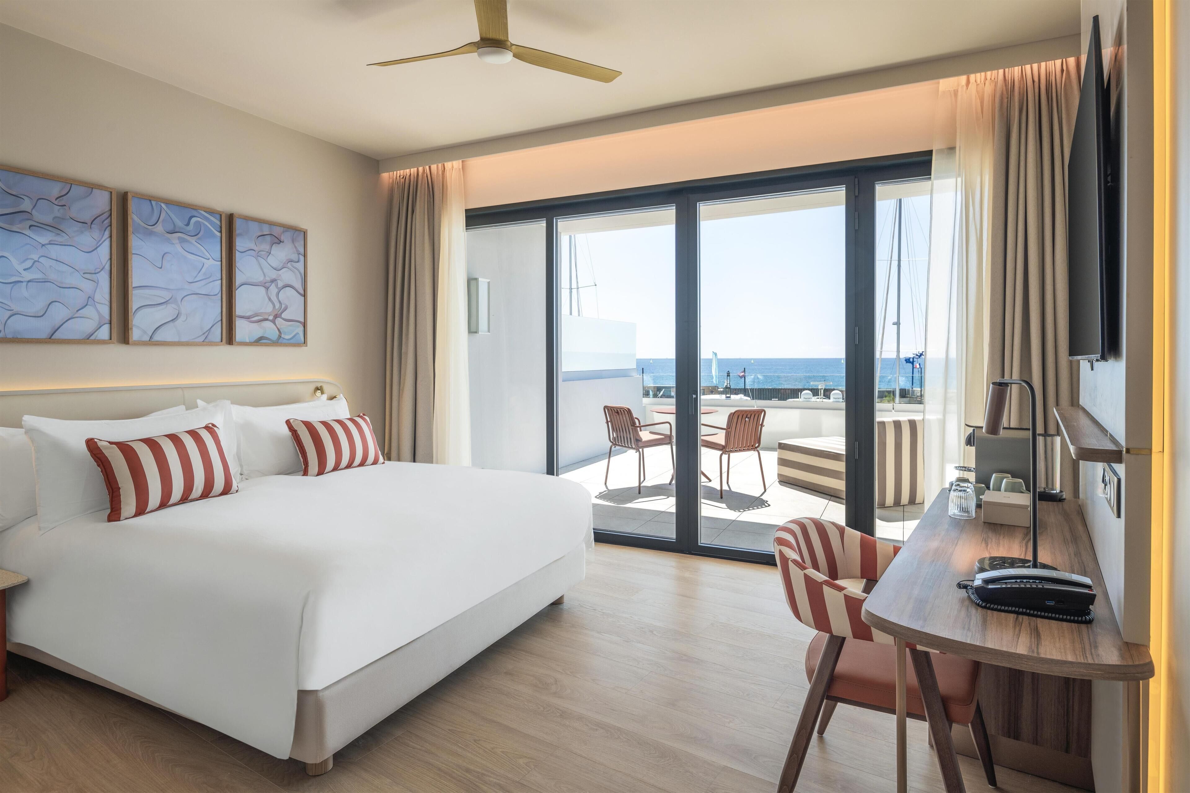 A hotel room with a bed, a desk with a chair, a television, and a view of the sea through large sliding doors.