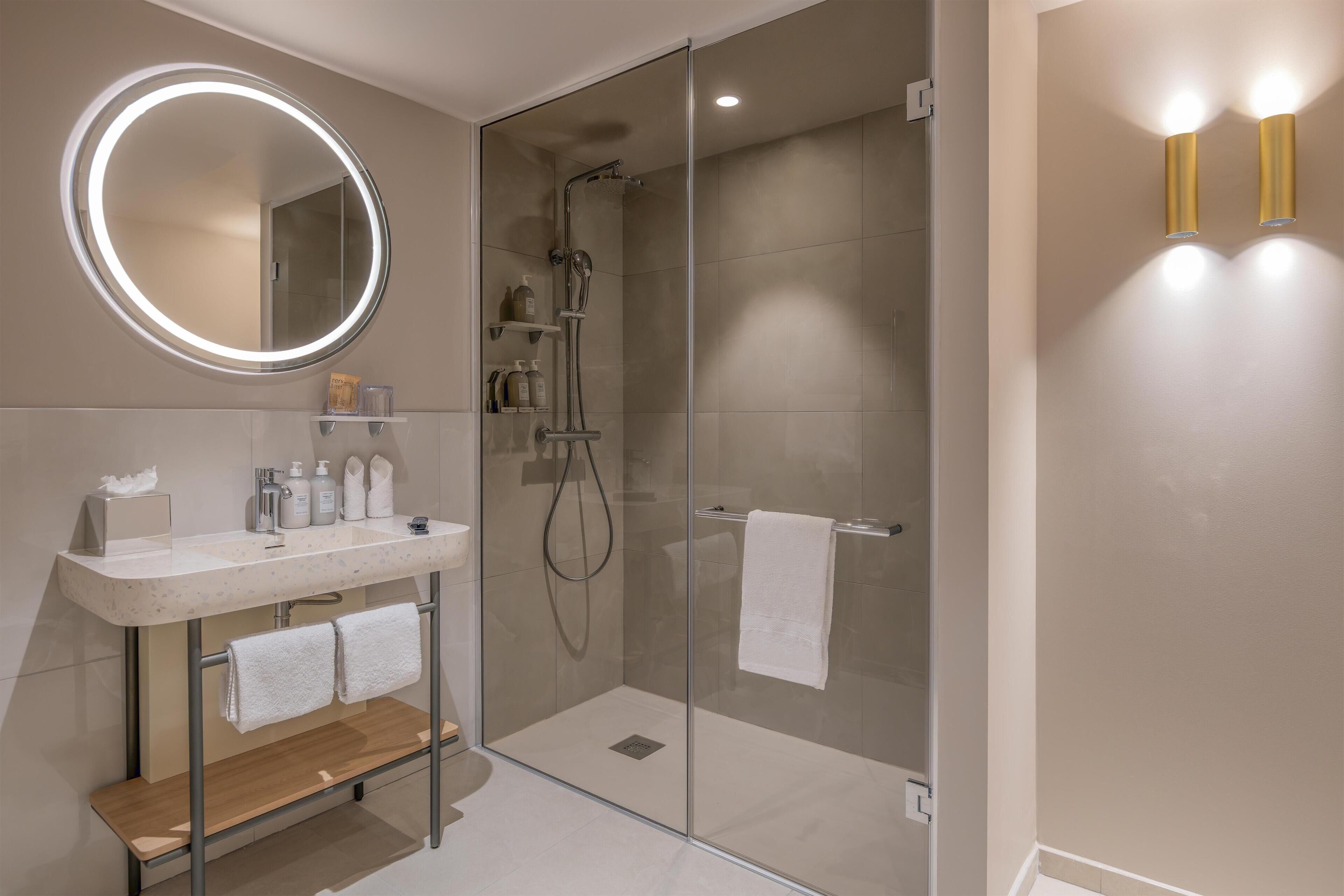A modern bathroom with a glass-enclosed shower, a round mirror with integrated lighting, and a vanity with a marble countertop.