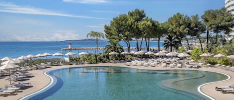 Una piscina de diseño curvo, rodeada de sillones de descanso y sombrillas, con vistas al mar y un faro a lo lejos.