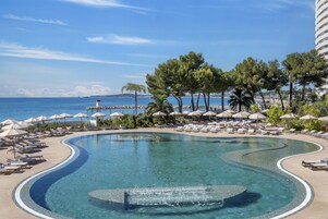 Una piscina de diseño curvo, rodeada de sillones de descanso y sombrillas, con vistas al mar y un faro a lo lejos.