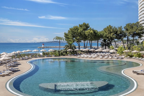Una piscina de diseño curvo, rodeada de sillones de descanso y sombrillas, con vistas al mar y un faro a lo lejos.