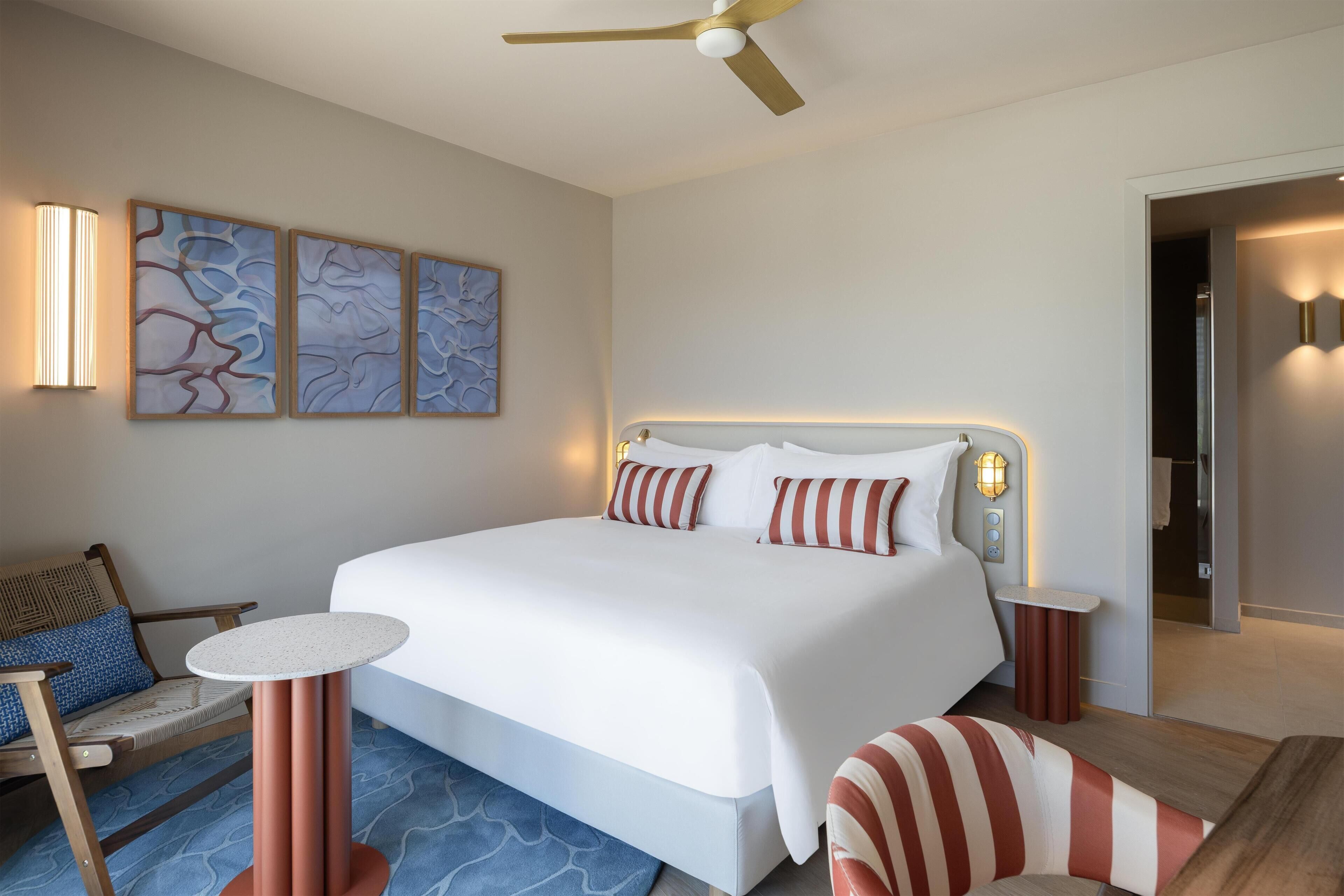 A hotel room with a large bed, bedside tables, a ceiling fan, wall-mounted artwork, and a seating area with a chair and a small table.