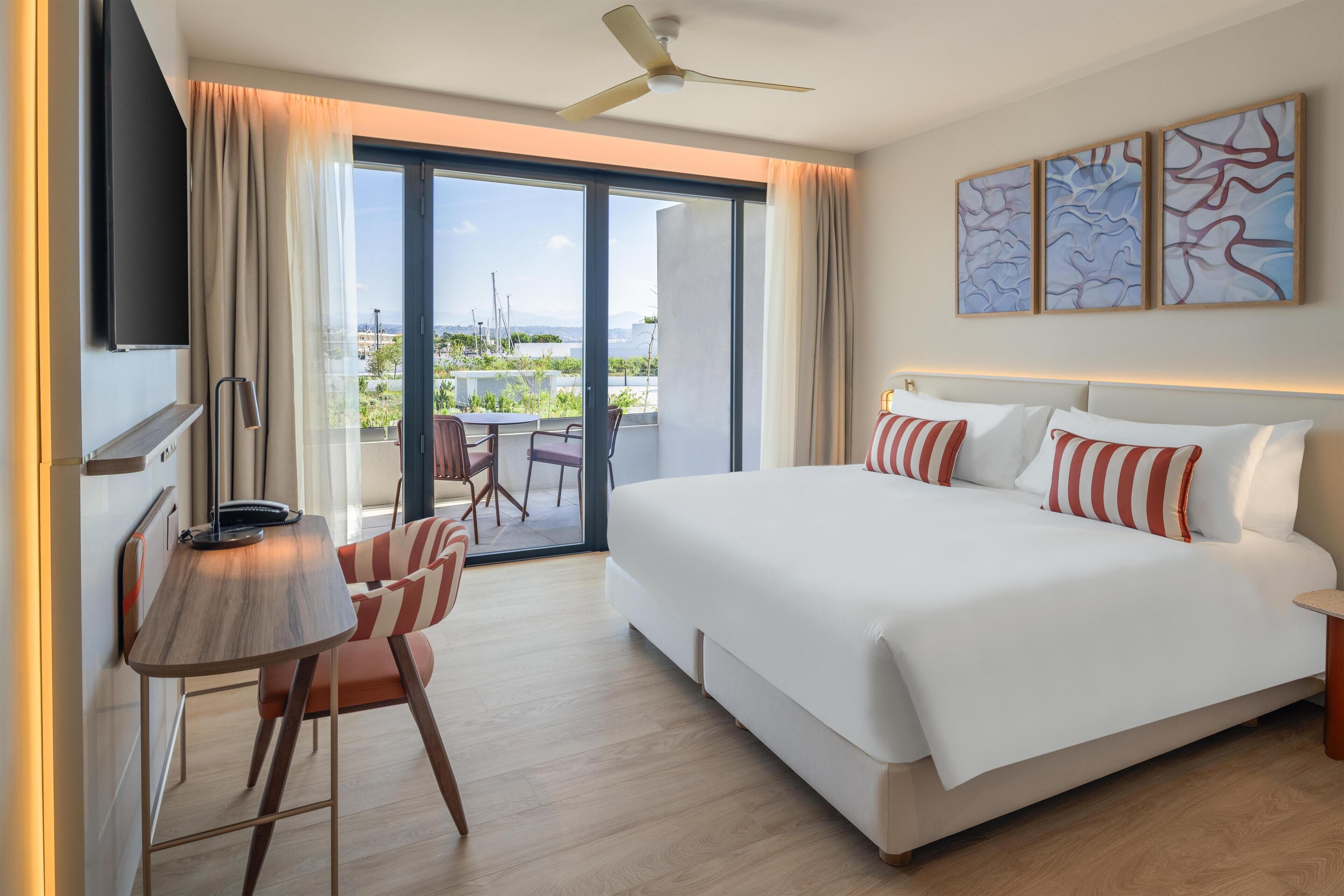 Habitación de hotel con una cama grande, un escritorio con silla, balcón con vistas y obras de arte en la pared.