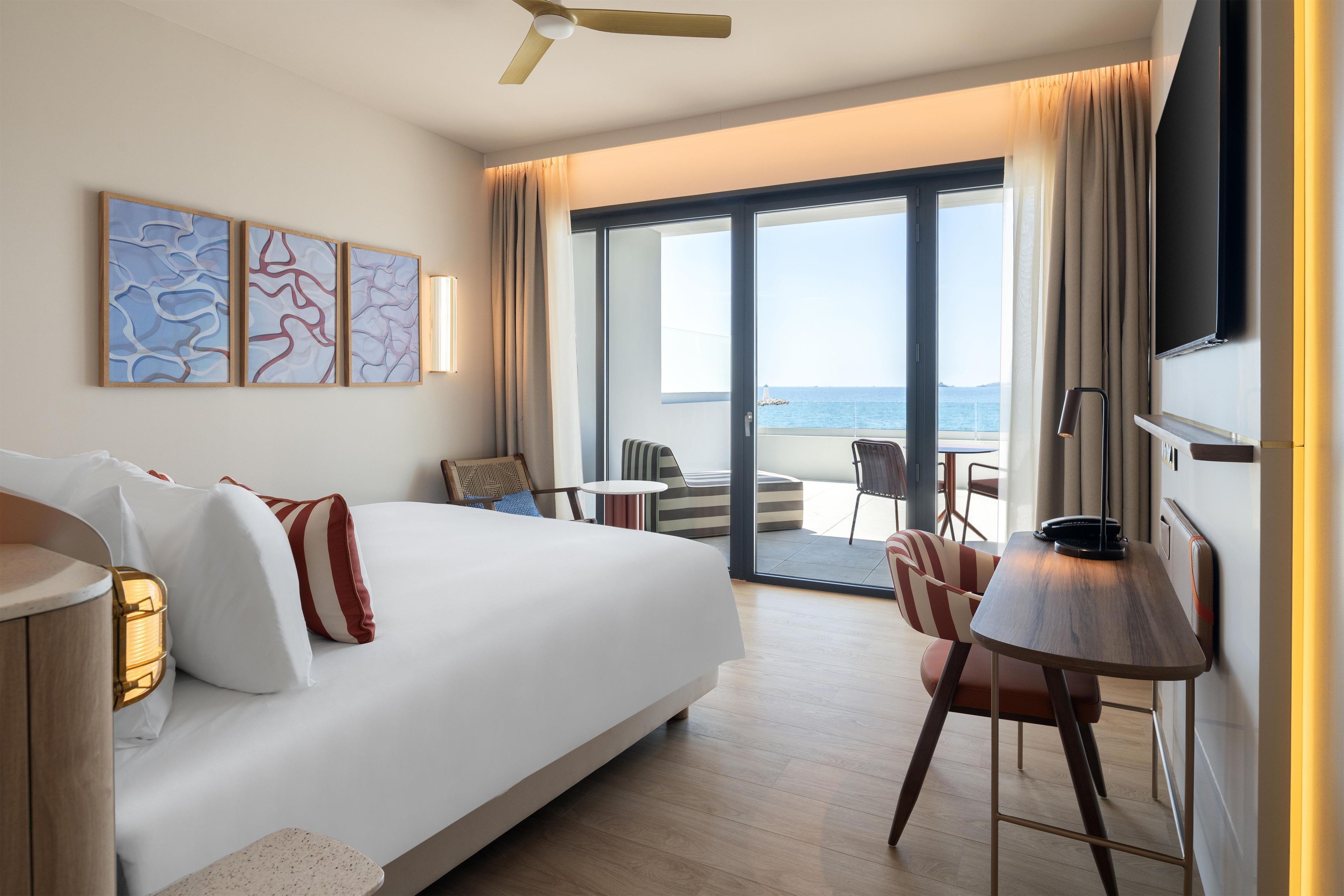 A hotel room with a bed, a desk, a chair, a TV, and a view of the sea.