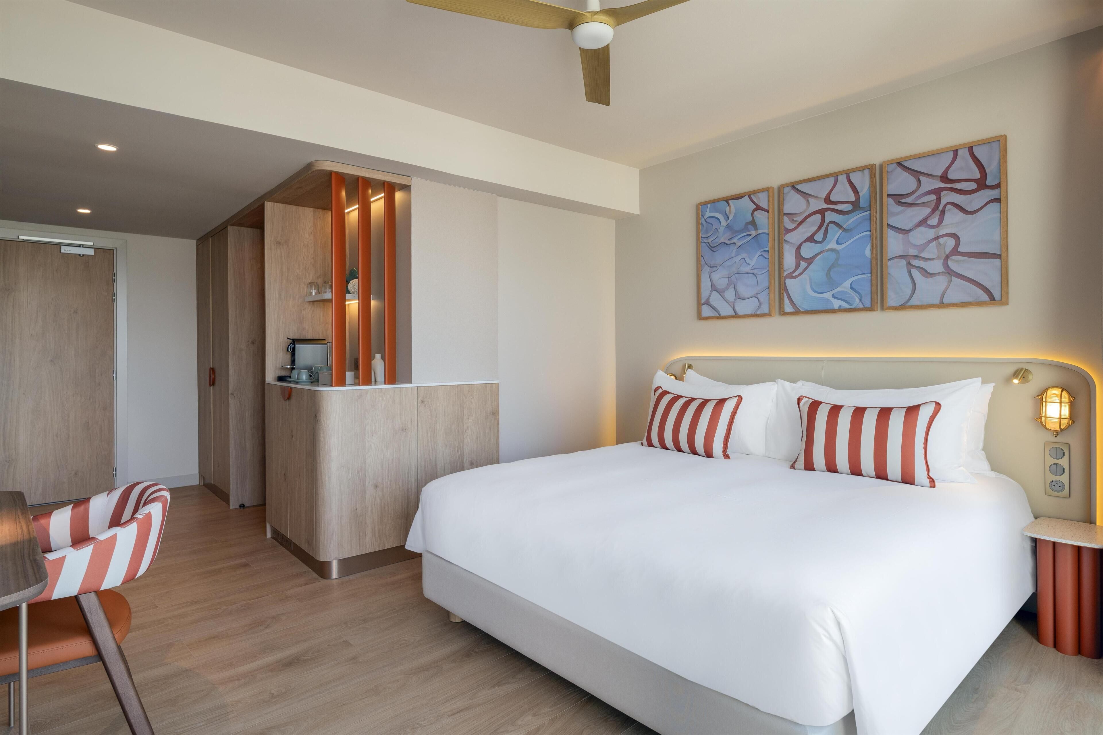 Habitación de hotel con una cama grande, un escritorio y una silla. Cuenta con ventilador de techo, una obra de arte en la pared y una mesita de noche con lámpara.