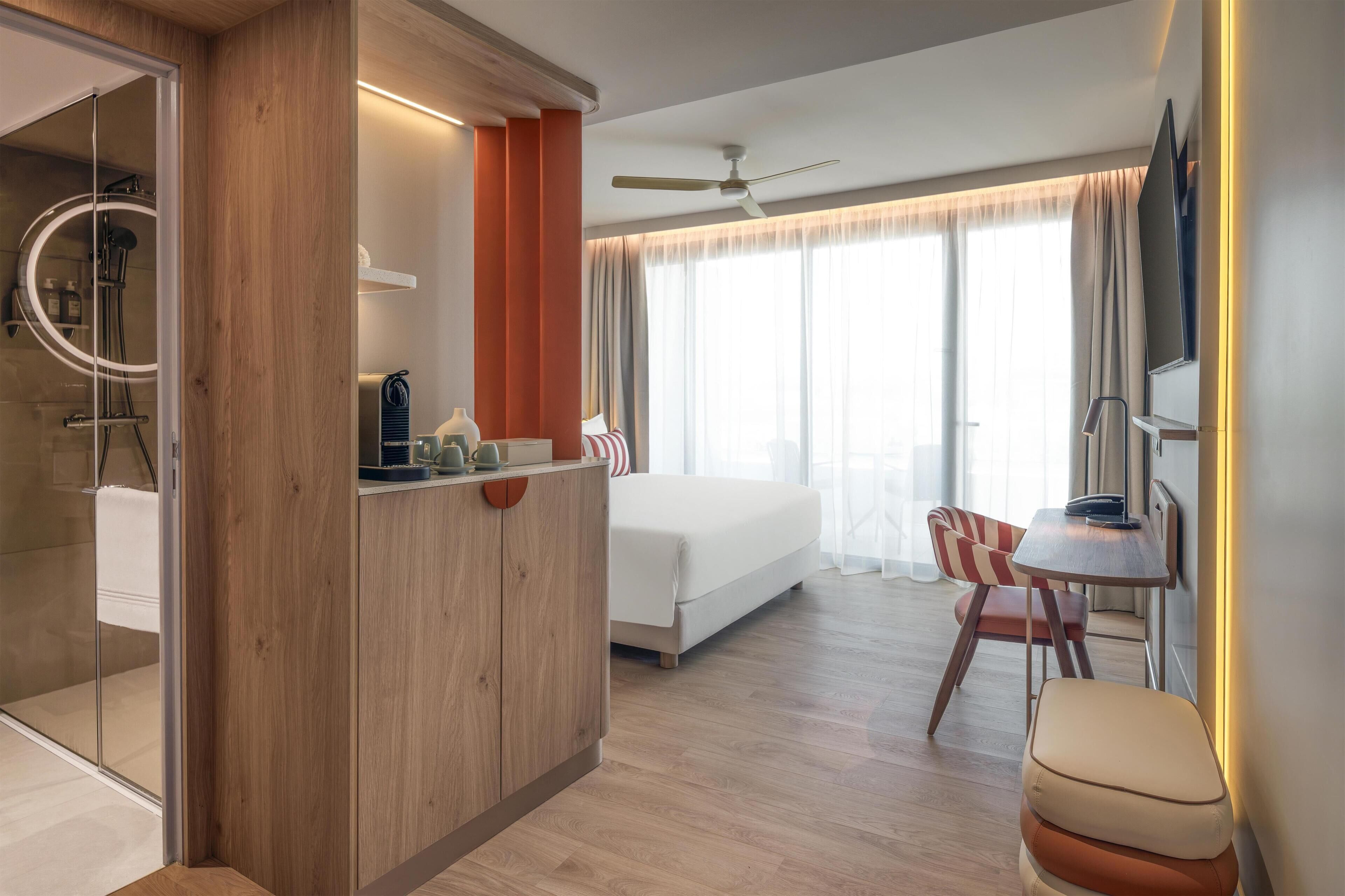 Una habitación de hotel moderna con cama, escritorio, silla, zona de ducha y un amplio ventanal con cortinas.