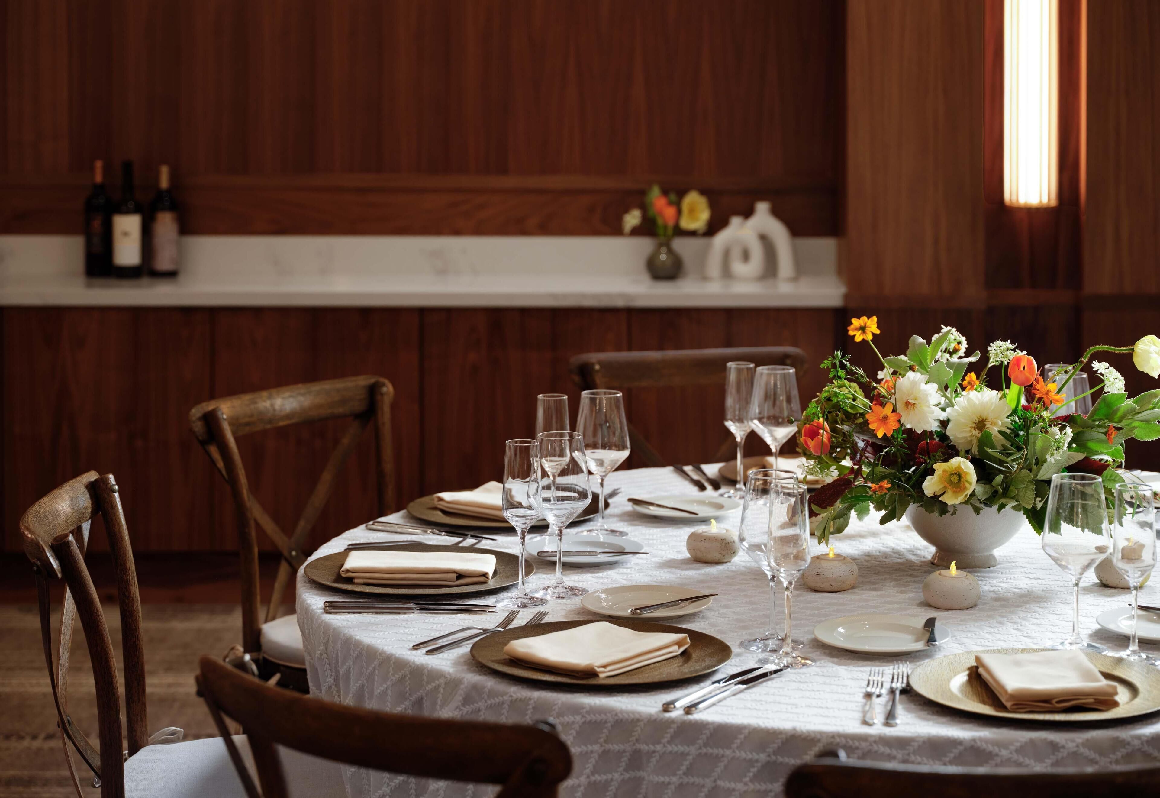 Mesa de comedor dispuesta con mantel blanco, platos, cubiertos y un centro de mesa floral.